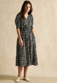 Schwarzes Blumenkleid mit kurzen Puffärmeln, geraffter Taille und knielangem Rock. Verfügt über weiße Blumenmuster und Taschen. Getragen mit weißen Slippern.