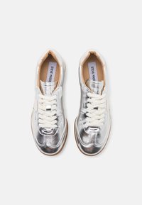 Metaliczne srebrne sneakersy o gładkiej fakturze, białe sznurówki i wyściółka w odcieniu brązowym. Posiadają okrągły czubek i przeszycia.