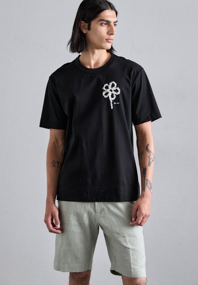 Schwarzes Baumwoll-T-Shirt mit einem strukturierten weißen Blumenmuster auf der linken Brust, kombiniert mit hellgrünen Leinen-Shorts.