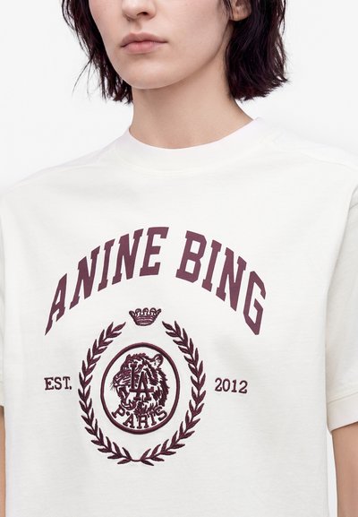 Νεαρό άτομο που φοράει άσπρο T-shirt με μαρονού κόκκινο κείμενο "ANINE BING,", έμβλημα τίγρης, στεφάνι δάφνης, κορώνα και σχέδιο "EST. 2012 PARIS".