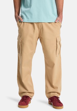 Person trägt beige Cargohose mit mehreren Seitentaschen, hellblaues Hemd und rote Sneaker mit Gummisohlen, steht mit den Händen in den Taschen.