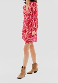 Femme portant une robe portefeuille rouge à fleurs avec manches longues et des bottines en daim beige, debout devant un fond clair uni.