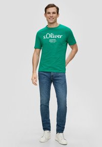 Groene katoenen T-shirt met witte tekst, "s.Oliver ELEVATED ESSENTIALS SINCE 1969," gecombineerd met blauwe spijkerbroeken en witte sneakers.
