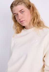 Sweat-shirt couleur crème en matériau doux, avec des poignets et un col côtelés, portant un petit logo blanc sur la poitrine gauche.