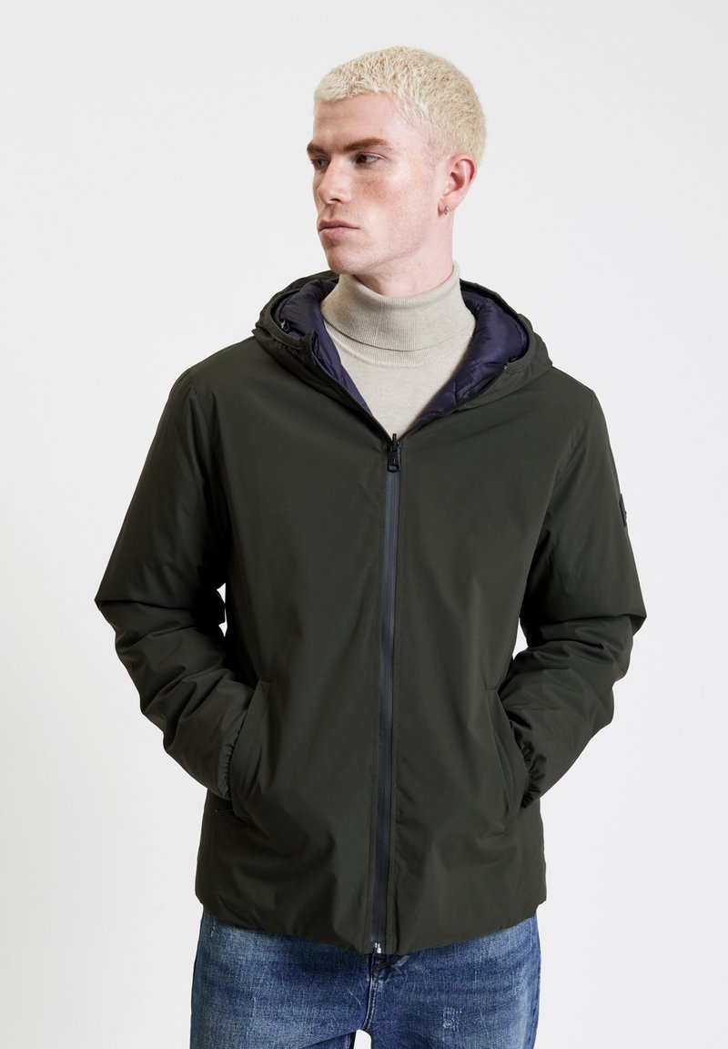 Giacca leggera con cappuccio di colore verde scuro, con zip, tasche laterali e una fodera interna scura in contrasto. Finitura texturizzata con un design moderno.
