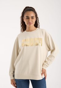 Beżowa bluza z długimi rękawami, z błyszczącą graficzną aplikacją logo w kolorze żółtym. Miękka tkanina o luźnym kroju i ściągaczach.