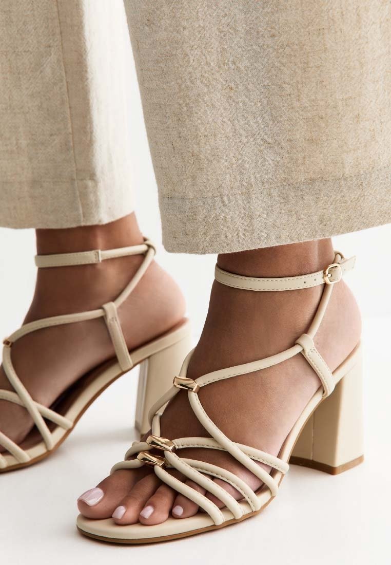 Pieds chaussés de sandales beiges à talons blocs avec fines lanières et accents dorés, associées à un pantalon large beige clair.