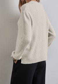 Pull côtelé beige clair en tissu tricoté texturé, avec manches longues et coupe décontractée, associé à un pantalon noir.