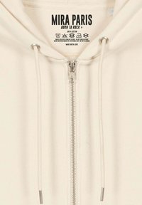 Sweat à capuche en coton crème avec une fermeture éclair à l'avant, des éléments en métal argenté et des cordons de serrage réglables. L'étiquette indique "MIRA PARIS, BORN TO ROCK."