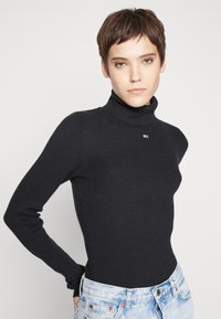 Maglione nero a coste con colletto a lupetto, maniche lunghe, design aderente e un piccolo logo sul petto. Abbinato a jeans in denim azzurro chiaro.