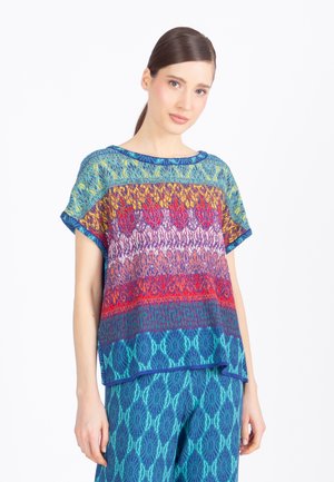 Donna con capelli lisci e scuri che indossa una maglietta a maniche corte multicolore con motivi e pantaloni blu con motivi, in piedi davanti a uno sfondo bianco neutro.