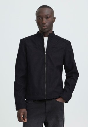 Man draagt een zwarte jas met rits over een wit shirt en donkere spijkerbroek, staat met één hand in de zak tegen een effen achtergrond.