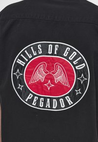 Black fabric with a circular patch showing white wings, a star, and text "Hills of Gold Pegador" in white on red and black background.