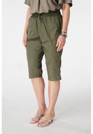 Vrouw die olijfgroene kniebroek draagt met elastische taille, trekkoord, zijzakken, een taupe top, armbanden, ringen en zilverkleurige sandalen met hak.