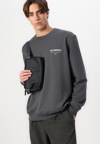 Felpa grigia con colletto rotondo e logo bianco "ALLSAINTS", abbinata a una borsa a tracolla nera con superficie textured e tracolla regolabile.