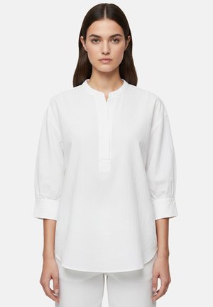 Donna con capelli castani lisci indossa una camicia bianca a maniche lunghe e morbida con una patta parziale di bottoni, in piedi davanti a uno sfondo semplice.