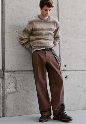Junger Mann trägt einen gestreiften Strickpullover und braune weite Hosen mit schwarzen Stiefeln, steht an einer Betonwand und schaut nach unten.