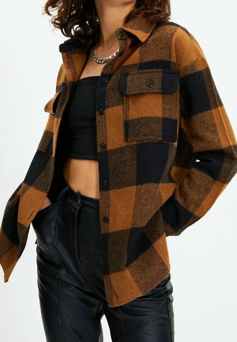 Femme portant une chemise à carreaux marron et noir boutonnée par-dessus un crop top noir et un pantalon en cuir noir, main dans la poche.