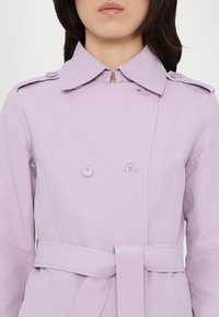 Lilla trench coat kakste erineva nupuga, suure krae ja vööga taljes. Omab kahte nuppu ja peeneid õmblusdetailide.