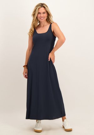 Vrouw met lang blond haar die een mouwloze marineblauwe maxi-jurk en witte sneakers draagt, glimlachend met één hand in haar zak.