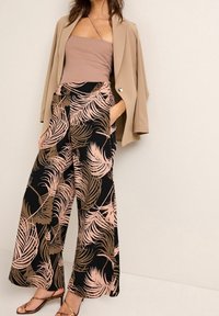 Beige blazer over een taupe top, gecombineerd met wijde, zwarte broeken met roze en bruine palmbladerenpatronen. Bruine sandalen met banden.