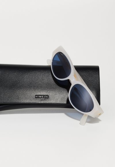 White cat-eye sunglasses with dark lenses, resting on a textured black case featuring a silver logo. Simple and modern design.