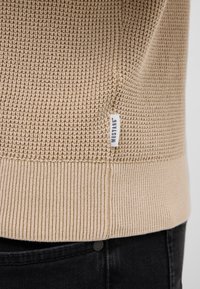 Beżowy sweter dzianinowy z teksturowanym korpusem i ściągaczami. Posiada małą, białą metkę marki po boku, w połączeniu z czarnymi dżinsami.