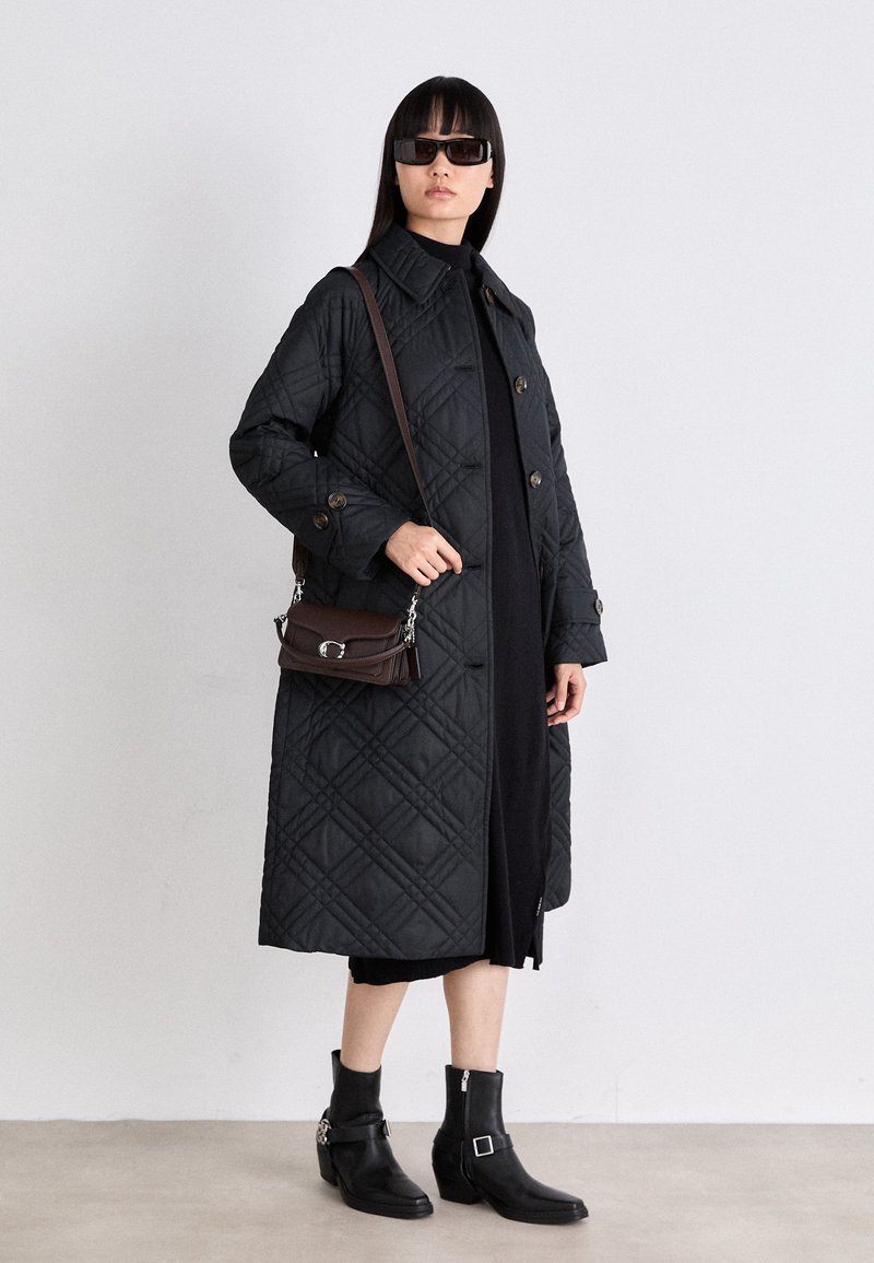 Cappotto lungo nero trapuntato con grandi bottoni, tasche laterali e colletto. Abbinato a un dolcevita nero, una borsa a spalla marrone e stivaletti neri.