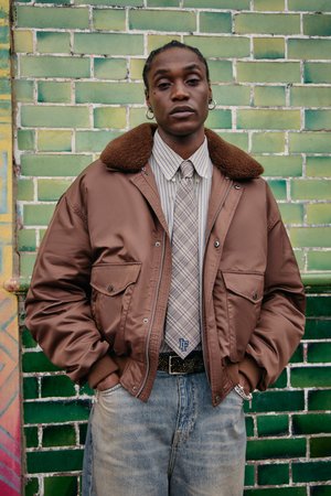 Jeune personne portant une veste bomber marron avec col en fourrure, chemise à carreaux et cravate, se tenant les mains dans les poches contre un mur en carrelage vert.