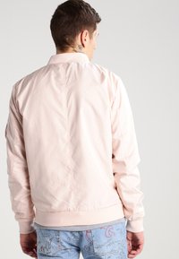 Lys rosa bomberjakke med en glatt, blank overflate; har ribbede ermer og linning; enkelt design uten synlig utstyr eller mønstre.