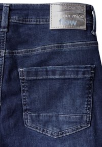 Tummansiniset farkut, joissa on takatasku ja hopeakirjaiminen vyötärömerkki. Kangas on sileä ja siinä on häilyvää kulumista.