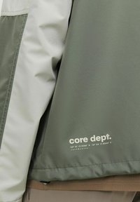 Grøn og lys grå jakke med en glat tekstur, prydet med "core dept." trykt i hvid, samt koordinater under logoet.