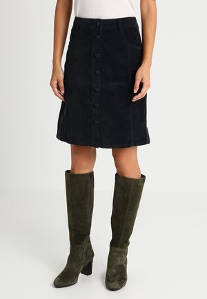 Femme portant une jupe en velours côtelé noire à boutons sur le devant, un haut blanc et des bottes à talons en daim vert olive montant jusqu'aux genoux, posant devant un fond uni.