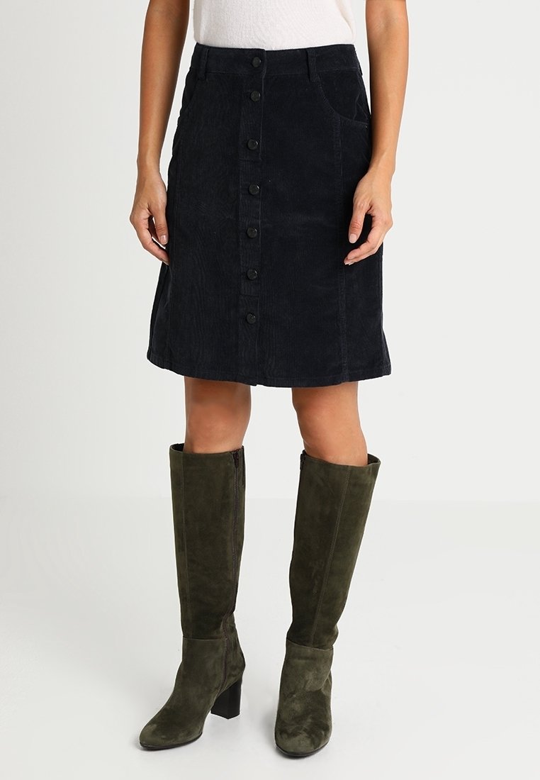 Femme portant une jupe en velours côtelé noire à boutons sur le devant, un haut blanc et des bottes à talons en daim vert olive montant jusqu'aux genoux, posant devant un fond uni.