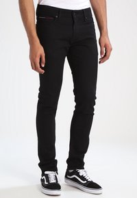 Black denim jeans with a slim fit, featuring five pockets and subtle red stitching detail at the front, paired with black and white sneakers.