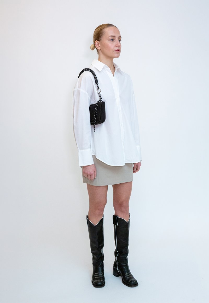 Γυναίκα που φοράει ένα λευκό oversized πουκάμισο, μπεζ μίνι φούστα, μαύρα μπότες μέχρι το γόνατο και κρατάει μια μικρή μαύρη τσάντα στον ώμο.