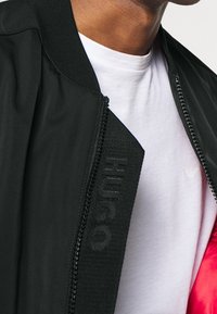 Blouson aviateur noir avec fermeture éclair et col côtelé, arborant le logo "HUGO". Porté sur un t-shirt blanc, mettant en valeur un design élégant.