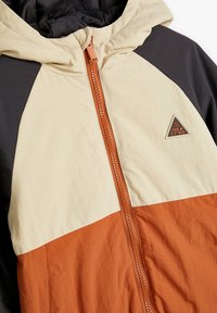 Kleurblokjack met paneel in beige, donkergrijs en oranje. Textuurstof met een rits aan de voorkant en een driehoekige logopatch.