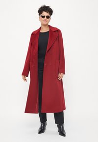 Casaco vermelho de dois botões com gola larga, grandes bolsos e textura suave, combinado com top e calças pretas, botas pretas brilhantes e óculos de sol.