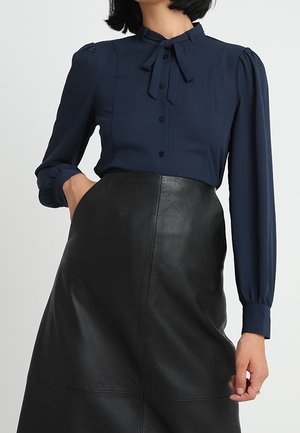 Mujer vestida con una blusa azul marino de manga larga con lazada en el cuello metida dentro de una falda de cuero negra de talle alto, mano en el bolsillo.