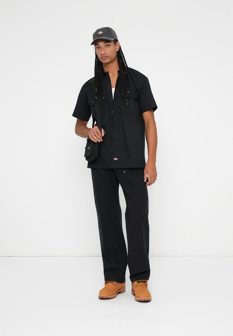 Chemise noire à manches courtes et pantalon, tous deux à coupe décontractée. Bottes marron avec lacets, portant un petit sac à bandoulière noir. La casquette est gris.