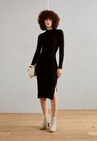 Czarna dzianinowa sukienka z prążkowanej tkaniny, z długimi rękawami i bocznymi rozcięciami, zestawiona z jasnoszarymi, teksturowanymi botkami za kostkę oraz kremową torebką.