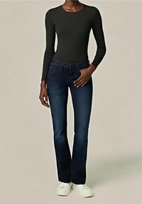 Femme portant un haut noir ajusté à manches longues, un jean bootcut bleu foncé et des baskets blanches, debout devant un fond uni.