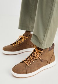 Zapatillas altas marrones hechas de material texturizado, con cordones naranjas, suela blanca y detalles de cuero marrón. Combinadas con pantalones verdes.