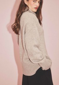 Helles beige Oversized-Pullover mit hohem Kragen, gerippten Bündchen und Saum. Weiche Textur, seitliche Taschen und lockere Passform. Hintergrund ist pink.
