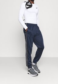 Pantalones de chándal azul marino de tela suave, con una franja lateral negra con logotipos blancos, combinados con zapatillas deportivas grises y una camiseta blanca ajustada.