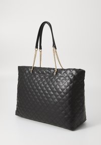 Sac fourre-tout en cuir, matelassé et noir, avec des poignées rehaussées de chaînes. Texture lisse et forme carrée structurée.
