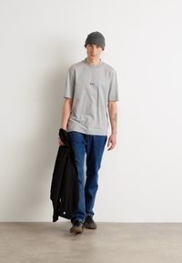 Graues Baumwoll-T-Shirt mit Logo, blaue Jeans, schwarze Jacke in der Hand, gestrickte graue Mütze und schwarze Schuhe. Einfaches, lässiges Outfit vor einer weißen Wand.