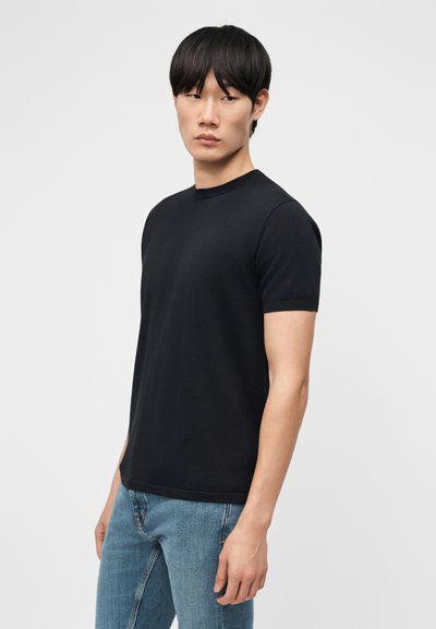 Mladý muž s čiernymi vlasmi, oblečený v jednofarebnom čiernom tričku s krátkym rukávom a modrých džínsoch, stojaci pred bielym pozadím.