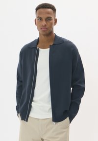 Marinblå zip-up kofta med nedvikt krage och långa ärmar, bärs över en ljus ribbad tröja. Slät textur och avslappnad passform.